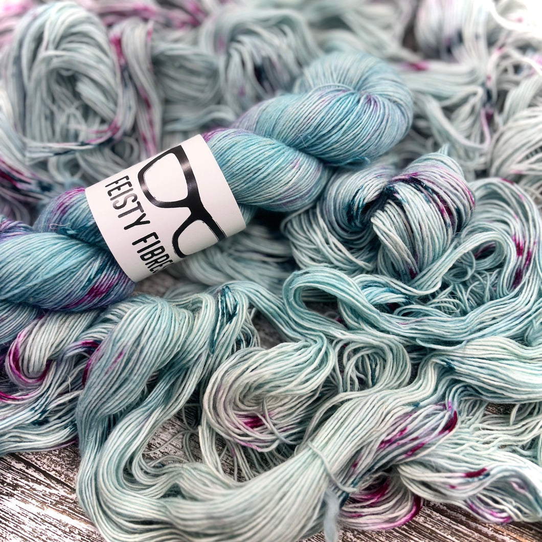 A skein of light teal blue merino linen yarn with  speckles of pink and purple a label that reads 'Feisty Fibres', surrounded by unspooled yarn.